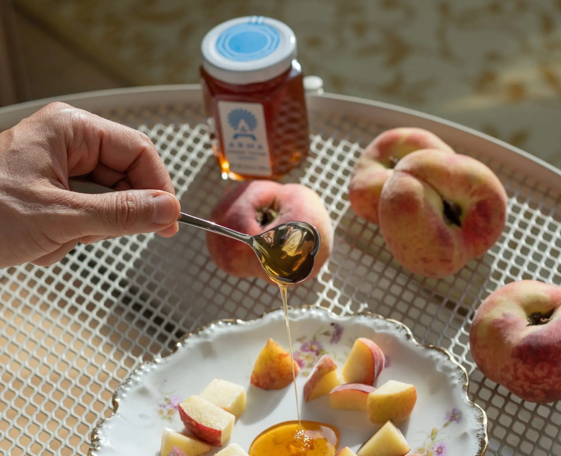 Greek Thyme Honey being drizzled over sliced peaches on a decorative plate with a jar of Arma honey in the background.