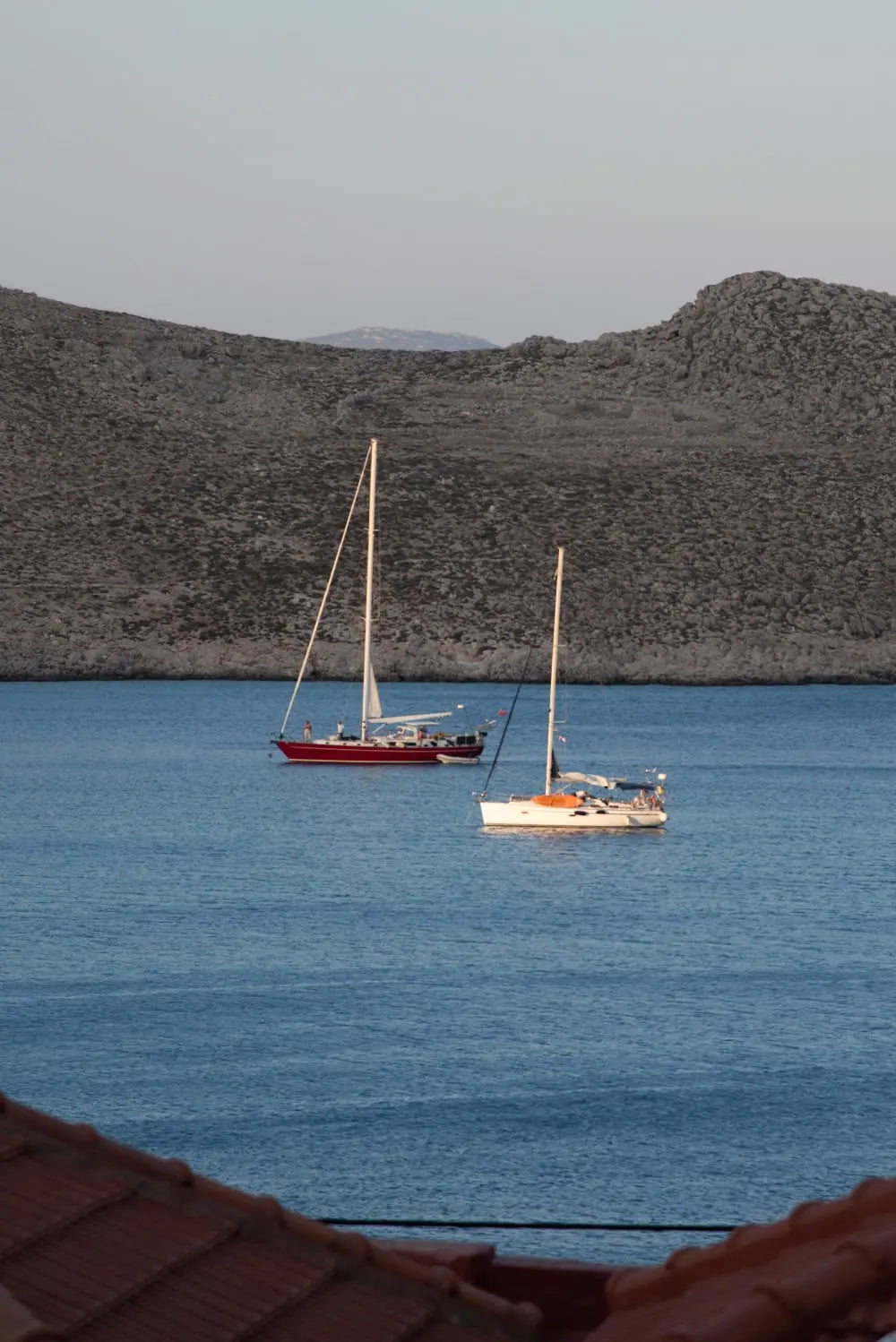 Two sailboats anchored in a calm blue sea with a mountainous Greek island in the background.