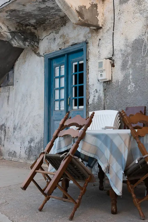 Outdoor setting with wooden chairs and a table covered with a striped tablecloth against a rustic Greek style building.